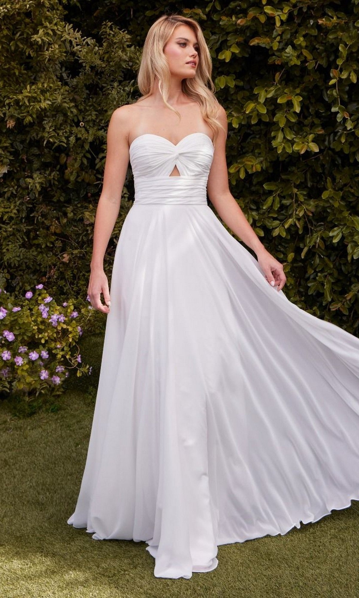 A woman wears the Strapless Long White Prom Gown: Ladivine 7435W, featuring a sweetheart bodice and small front cutout, as she stands outdoors on grass near green bushes and purple flowers.